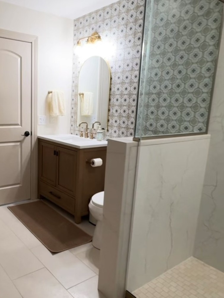Remodeled bathroom vanity area in Montgomery, IL by NSG Construction LLC featuring a walnut vanity with white countertop, brushed gold bridge faucet, arched gold mirror, brass vanity light, and floor-to-ceiling decorative patterned tile accent wall