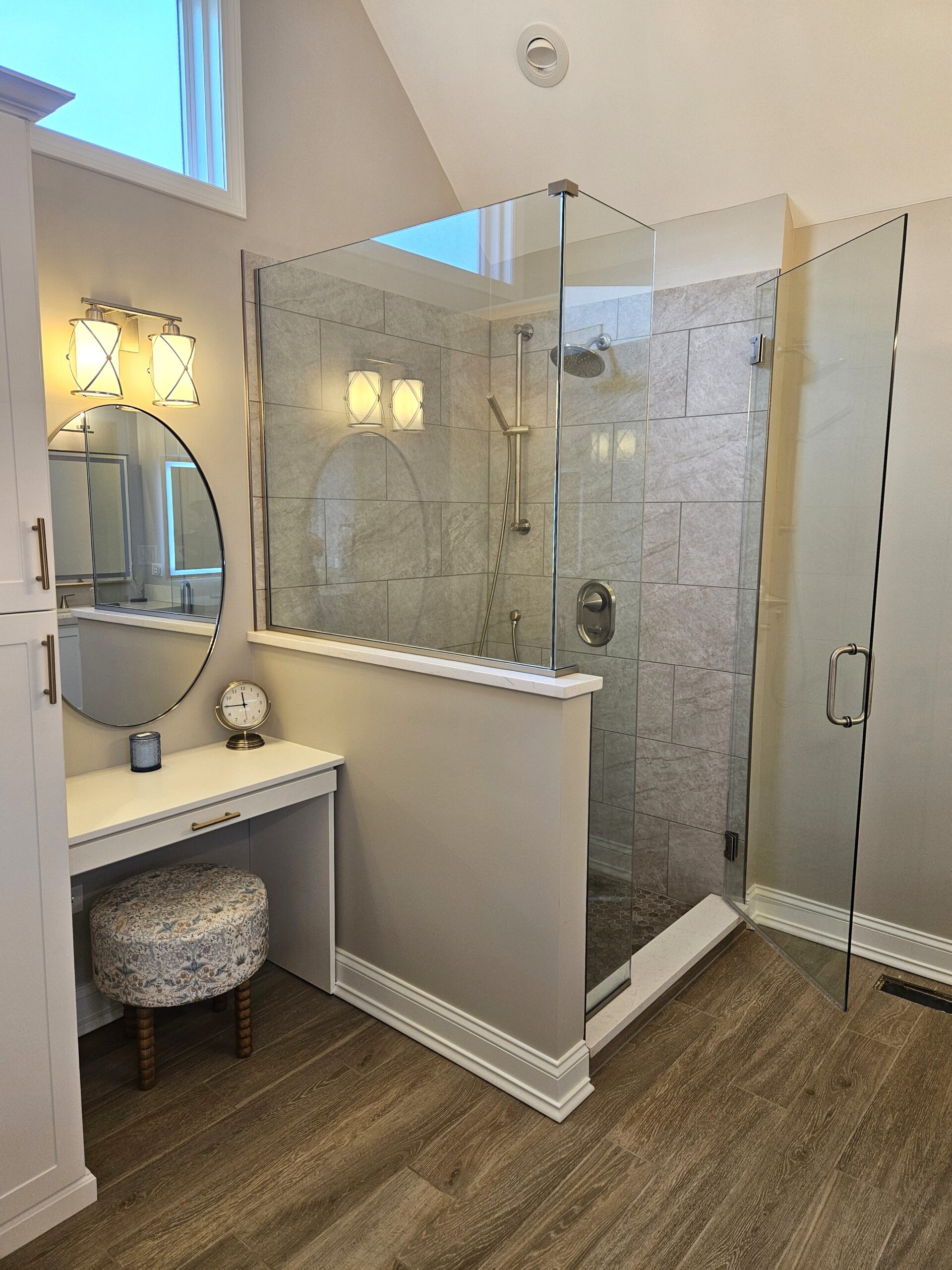 Remodeled bathroom in Naperville, IL by NSG Construction LLC featuring a custom gray tile walk-in shower with frameless glass enclosure, brushed nickel rain showerhead, wood-look tile flooring, and a built-in white makeup vanity with oval mirror