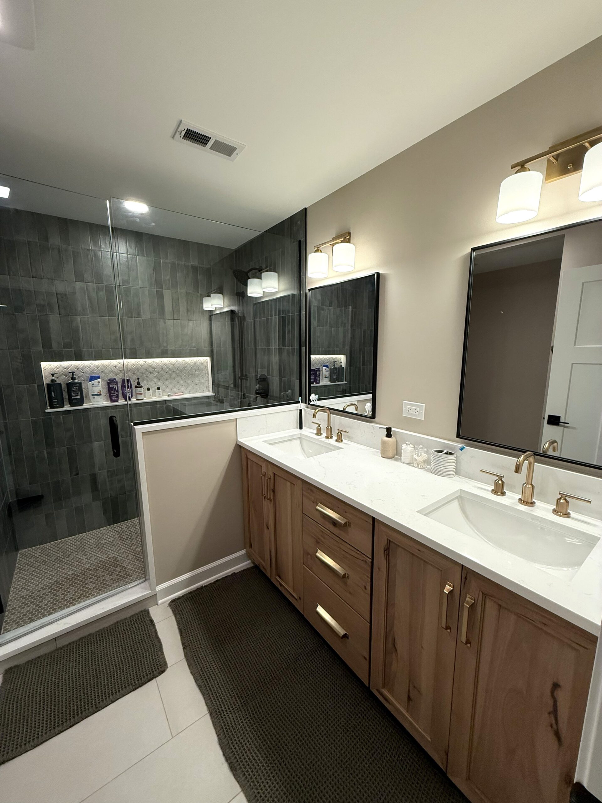 Bathroom addition in Somonauk, IL by NSG Construction LLC featuring custom wood double vanity with quartz countertop, dual undermount sinks, brushed gold faucets, gold vanity lights, black framed mirrors, and a custom green tile walk-in shower with glass enclosure and lighted built-in niche
